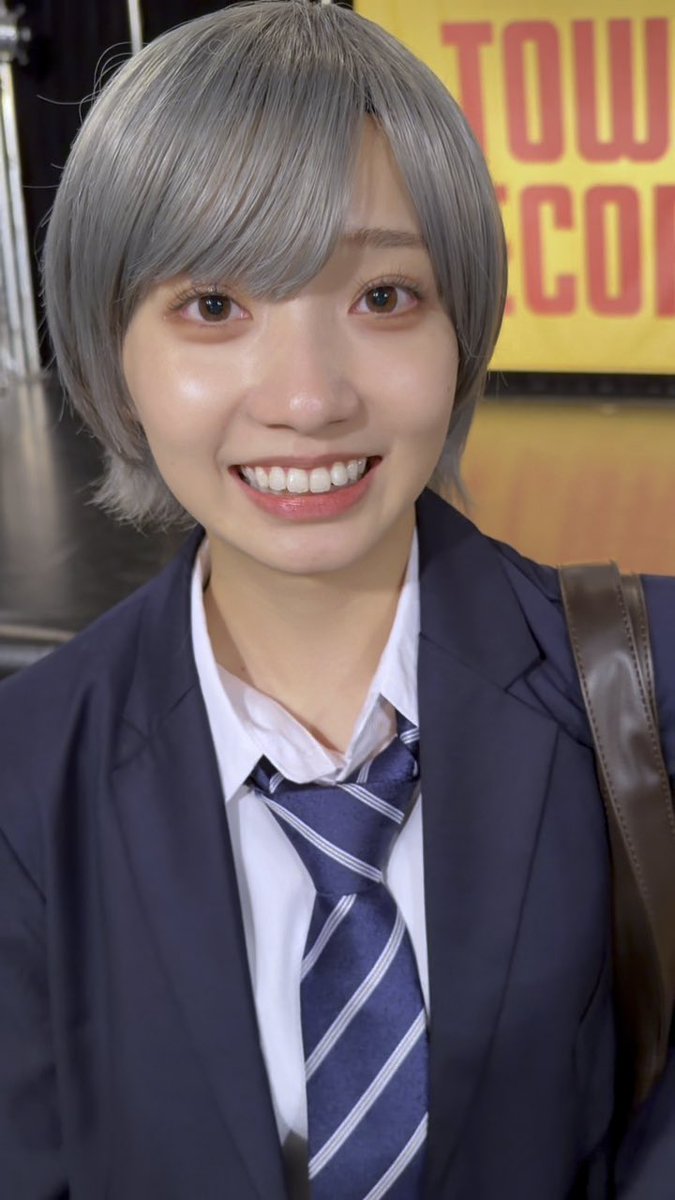 A young woman with short silver-gray hair smiles brightly at the camera, wearing a navy blazer, white collared shirt, and a blue-and-white striped tie, with a partially visible yellow 'TOW... COM' sign in the background.