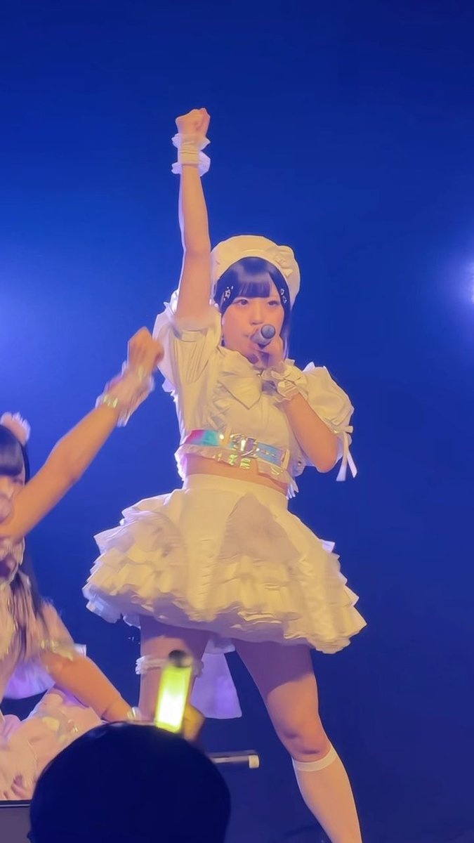 A Japanese idol performer in a frilly white outfit and headband sings into a microphone on stage with her fist raised, while another performer is partially visible on the left.
