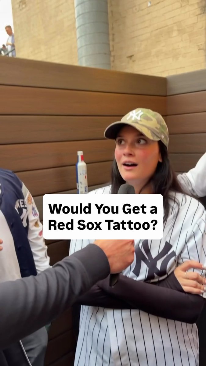 A woman wearing a New York Yankees pinstripe jersey and camo Yankees cap is being interviewed at what appears to be a baseball game, with a text overlay reading "Would You Get a Red Sox Tattoo?"—prompting her shocked reaction that matches the post's "BETWEEN THE WHAT????" sentiment, as the idea of getting a rival team's tattoo is unthinkable for a die-hard Yankees fan.