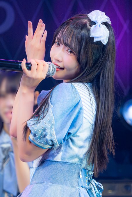 A young woman with a white hair bow performs on stage in a light blue outfit, singing into a microphone while holding her hand up.