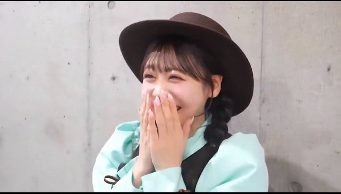 A young woman wearing a brown hat and light blue coat covers her mouth while laughing against a concrete wall.