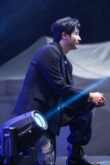 A young man in a black suit sits in profile on a stage illuminated by blue spotlights, appearing to hold a microphone during a performance.