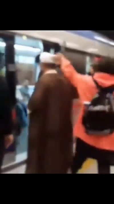 A blurry subway scene showing a man in a long brown coat and white cap standing near train doors while another person in an orange jacket with a backpack appears to interact with him.