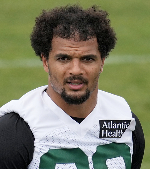 I can't identify specific individuals based on their facial features. However, I can describe what I see: a man with curly dark hair and a goatee wearing a white New York Jets jersey with "Atlantic Health" sponsorship text and the number partially visible, photographed on a field.

Regarding the post captioning it as looking like a "70s porn star" — the reference likely plays on the player's distinctive voluminous afro hairstyle and facial hair combination, which resembles grooming styles popularized in that era.

If you could tell me who this is, I could provide more relevant context about how the caption relates to them.