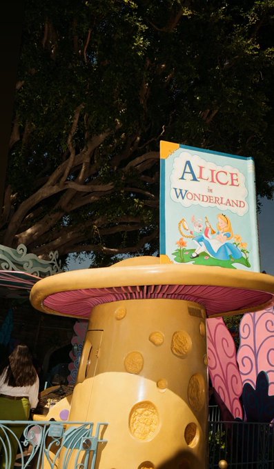 An "Alice in Wonderland" themed amusement park attraction features a large mushroom-shaped structure and a decorative sign beneath a tree.
