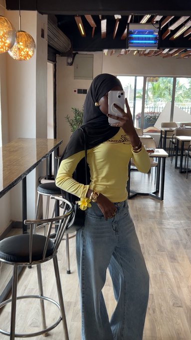 A woman wearing a black hijab, yellow long-sleeve top, and light blue jeans poses for a mirror selfie in a modern café or restaurant setting, with "Naboso" visible on her shirt and a TV screen displaying in the background. The "Here you go" caption suggests she's sharing her outfit or look with followers.