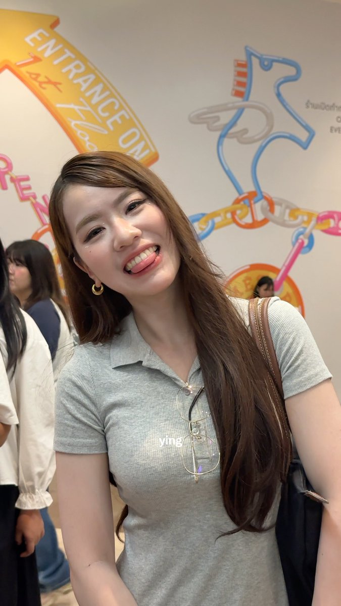 A smiling woman with long brown hair wearing a gray polo shirt stands in front of a colorful wall mural featuring the text "ENTRANCE ON" and "Jazz Kiss"; the image has a "ying" watermark.