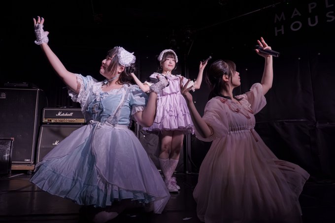 Three performers in elaborate pastel-colored maid-style dresses performing on stage in front of Marshall amplifiers with microphones in hand.