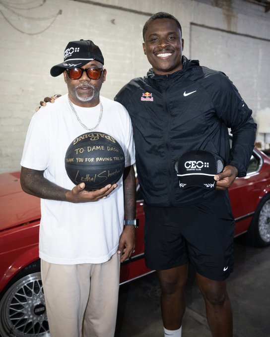 Two men posing together indoors with a red car in the background; the man on the left wears a black cap and sunglasses while holding a signed basketball that reads "TO DAME DASH THANK YOU FOR PAVING THE WAY" with LeShaun's autograph, and the man on the right wears a black Nike jacket and holds a "CEO" branded cap.