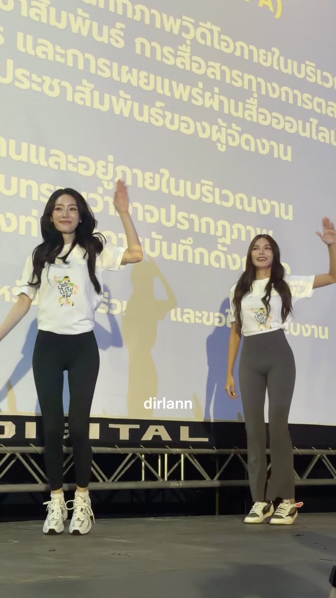 Two women in white tops and dark pants waving on stage in front of a Thai-language presentation screen, with "dirlann" watermarked on the image.