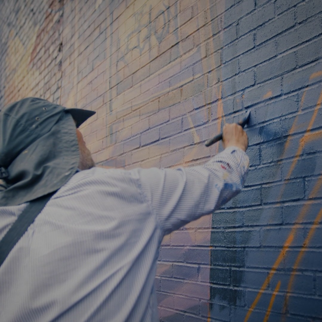 An artist in a bucket hat and striped shirt paints a brick wall mural in deep blue tones, with gold linework and a faded skull motif visible above, capturing the in-progress creation of the TYXNA project on Montreal's Boulevard Saint-Laurent.