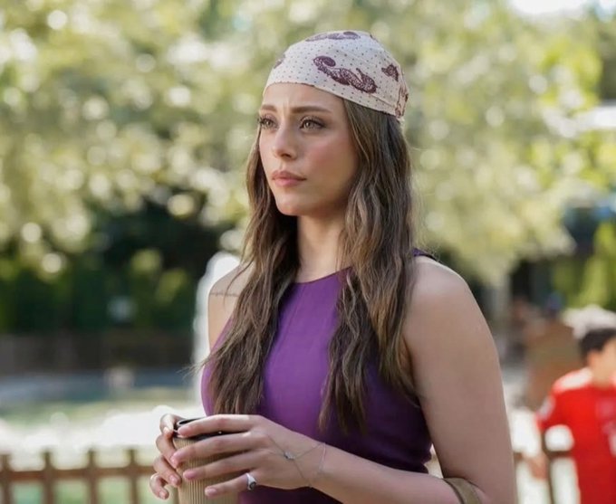 A woman with long wavy hair wearing a patterned bandana and a purple sleeveless top holds a coffee cup outdoors.