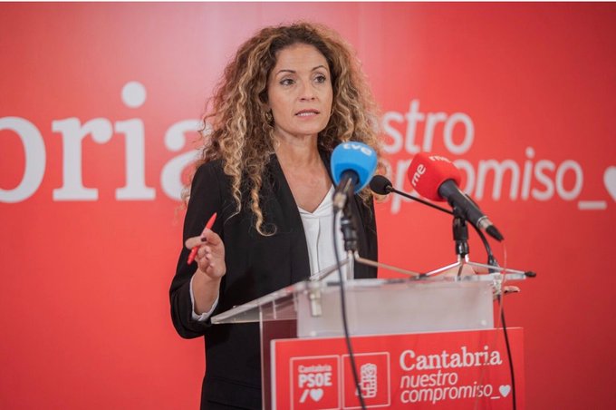 A woman with curly hair wearing a black blazer speaks at a podium during a press event for 'Cantabria nuestro compromiso' and PSOE, with 'tvnc' and 'rne' microphones visible.
