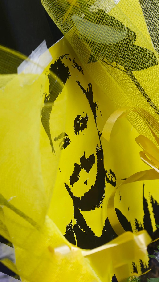 A black stencil-style portrait of a person's face surrounded by yellow tulle fabric and a gold ribbon.