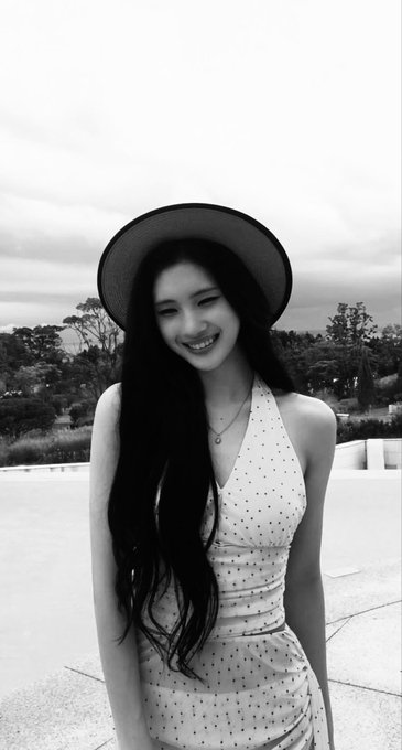 A black-and-white photograph of a smiling young woman with long dark hair, wearing a wide-brimmed hat and a polka-dot dress, standing outdoors.