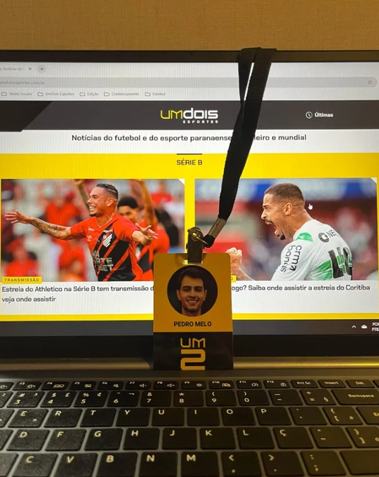 A lanyard with an ID badge labeled "PEDRO MELO" hangs over a laptop keyboard, with a web browser in the background displaying sports news from "umdois ESPORTES" about the Série B.