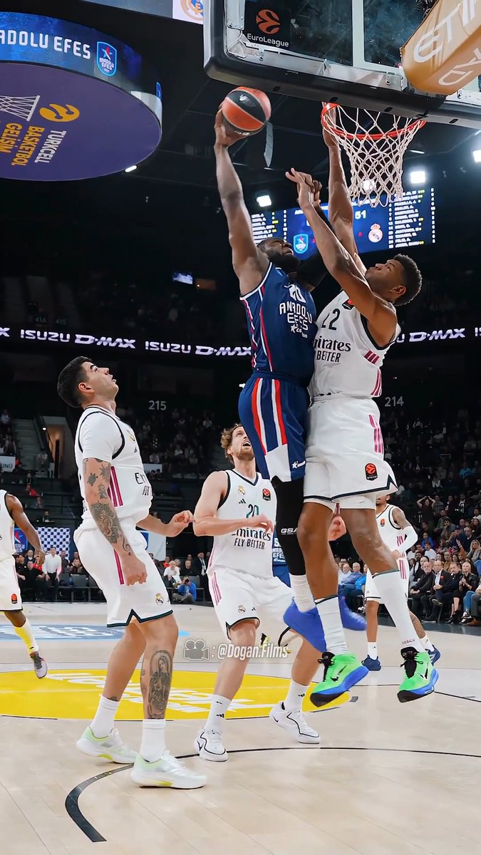 An Anadolu Efes basketball player in a blue jersey attempts a layup against Real Madrid players in white jerseys during a EuroLeague game, with watermarks '@dogan.films' and sponsor logos visible.