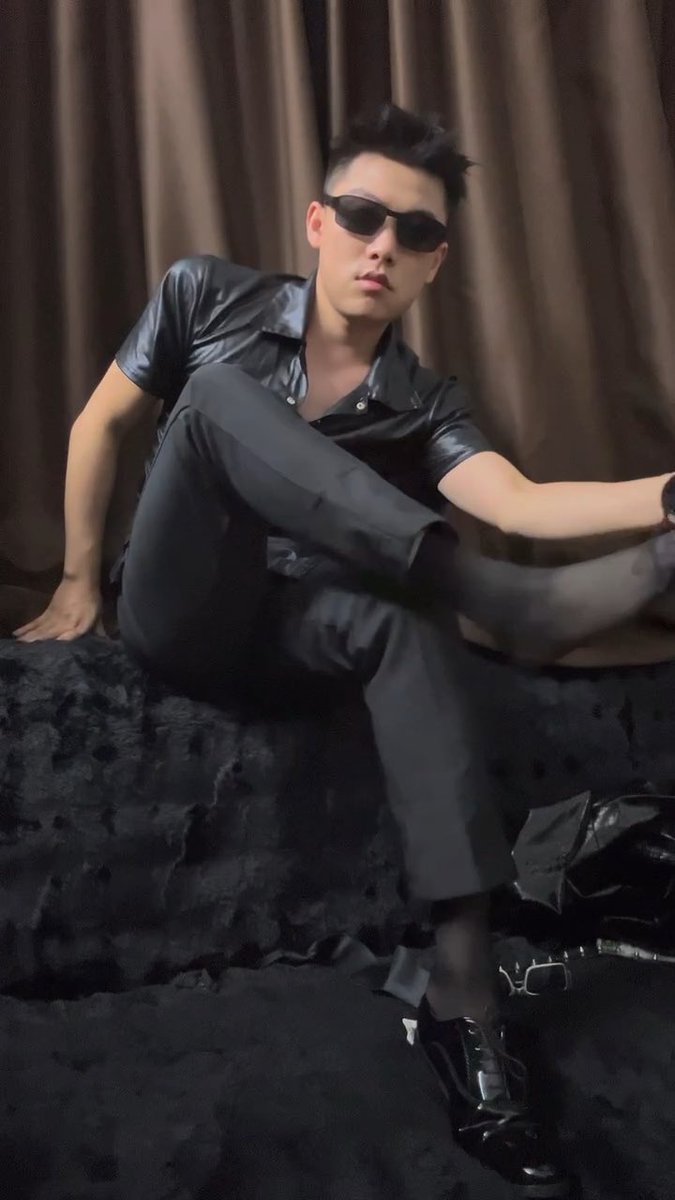 A young man with dark hair wearing black sunglasses, a black short-sleeved shirt, and dark pants reclines on a black surface with brown curtains in the background.