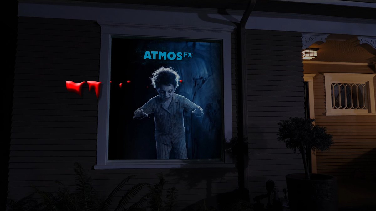 An ATMOS FX projection of a creepy doll in pajamas appears in a house window at night, likely as part of a Halloween decoration setup.