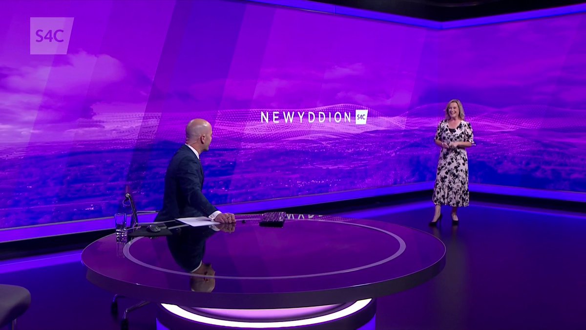 A brightly lit television news studio with purple and pink screens displaying 'NEWYDDION S4C,' featuring a man seated at a circular desk and a woman standing in a black and white floral dress.