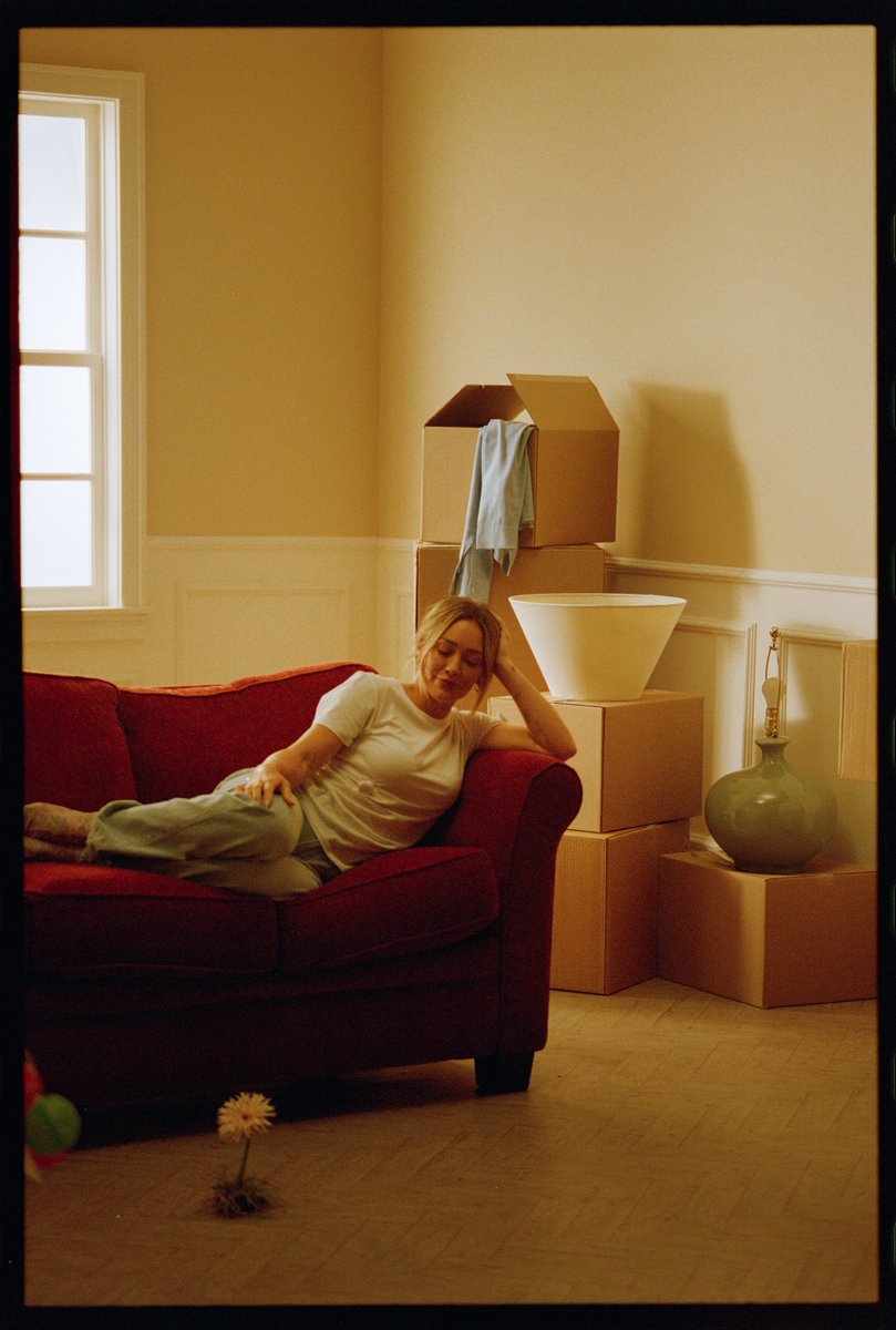 A blonde woman in a white shirt rests on a red couch in a yellow-walled room, surrounded by stacked cardboard moving boxes, a lamp, and a decorative vase.