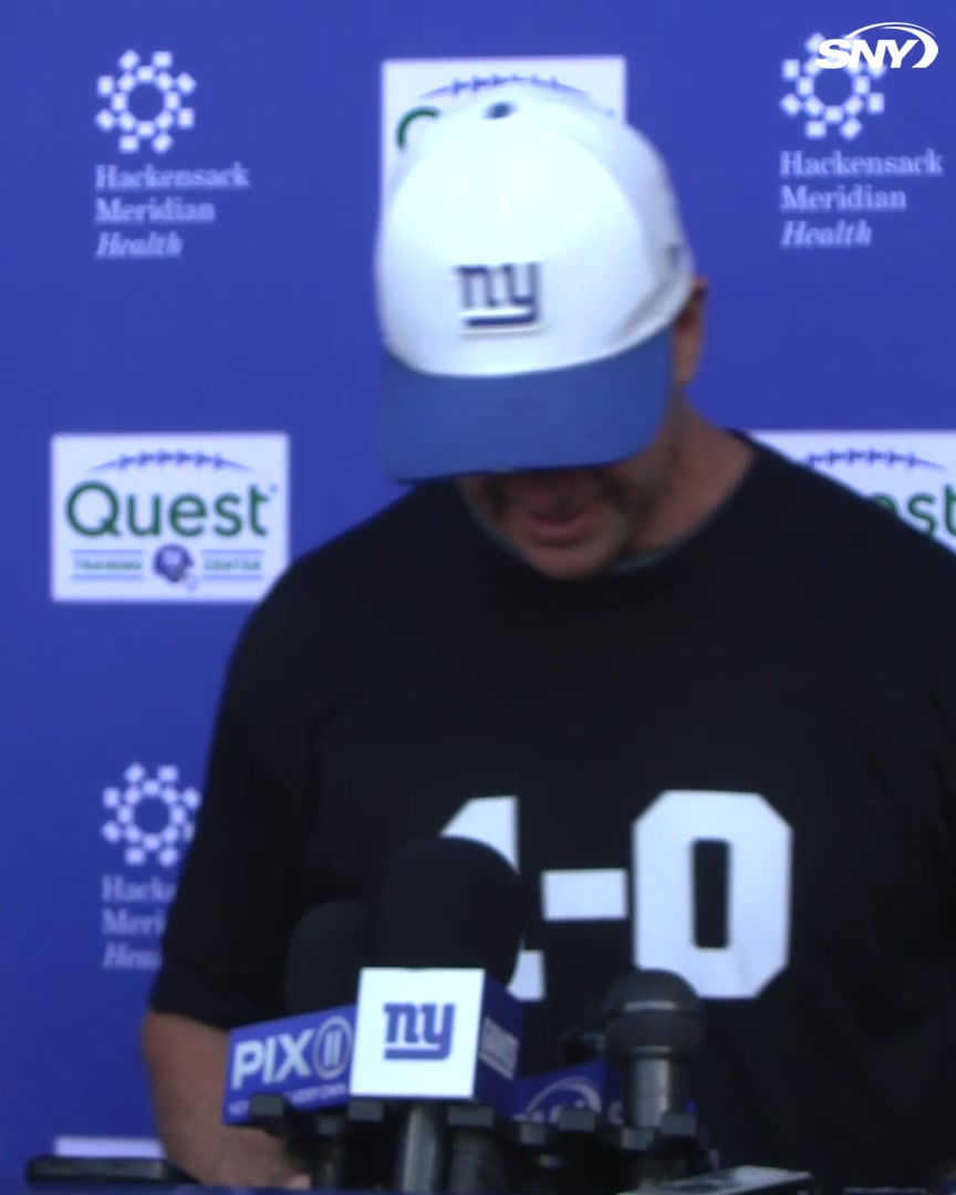 A person wearing a New York Giants cap and a "1-0" shirt speaks at a press conference podium with PIX11 and SNY microphones, while a backdrop displays Hackensack Meridian Health and Quest logos. The "1-0" shirt and Giants cap suggest a postgame press conference following an opening-week victory, with Harbaugh likely discussing the win before the Beatles comment was made.