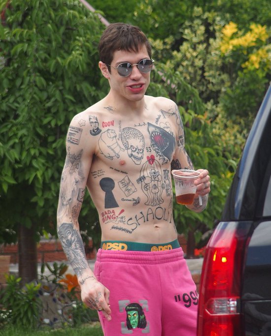 Pete Davidson stands shirtless outdoors, revealing the extensive tattoos he's currently in the process of removing, including visible text like "DADA BING!" and "CHAOL!" across his torso, paired with bright pink sweatpants as he holds a drink near an SUV.