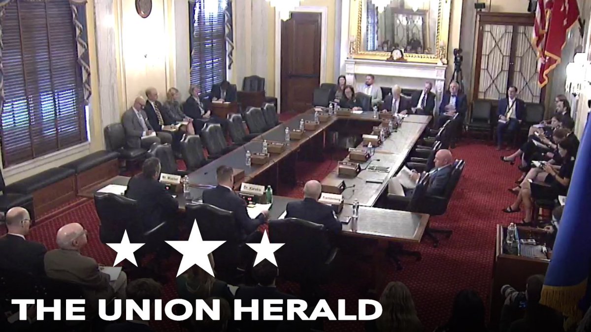 A wide shot of a formal government meeting in a wood-paneled room with people seated around a long table, overlaid with 'THE UNION HERALD' text and three stars.