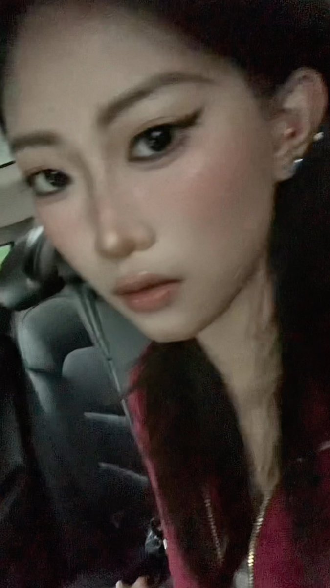 A close-up selfie of a woman with dark hair, reddish blush, and a maroon jacket sitting inside a car.