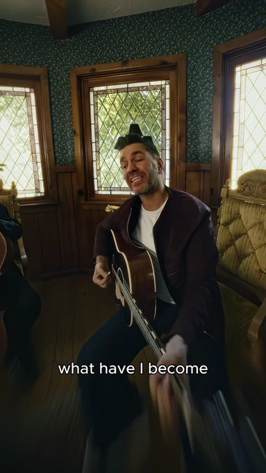 A man wearing a hat and maroon jacket plays an acoustic guitar in a wood-paneled room with leaded glass windows, accompanied by the caption "what have I become."