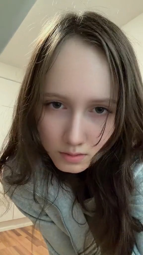 A close-up selfie of a young girl with long brown hair and blue eyes, wearing a light blue puffy jacket.