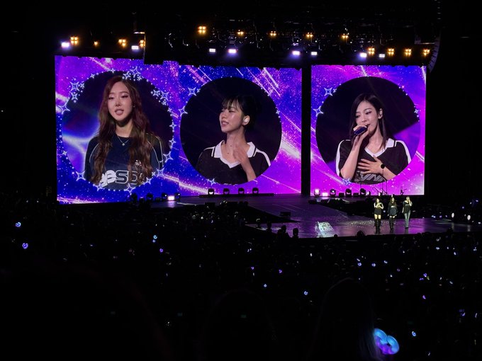 A concert stage features three large LED screens displaying portraits of K-pop performers against a purple cosmic background, with audience members holding glowing light sticks in the dark arena below.