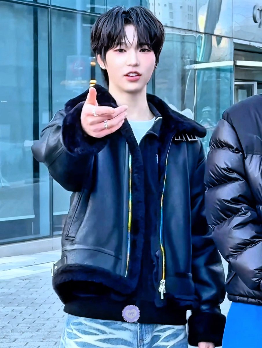 A young Korean male idol wearing a black leather jacket with fur trim over a light blue shirt, pointing at the camera, wearing multiple rings, photographed outside a glass building.