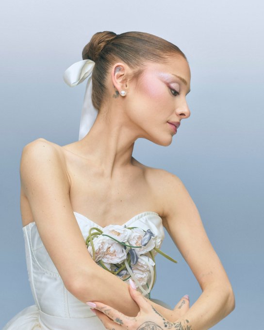 A woman with her hair in a bun and a white bow wears a strapless white dress, gazing downward at a bouquet of flowers she holds against a soft blue background.