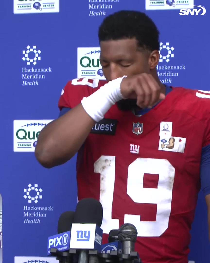 A New York Giants player wearing jersey number 19 wipes his face with a taped hand during a press conference, with "Hackensack Meridian Health," "Quest," "PIX11," and SNY logos visible on the backdrop and microphones. The image accompanies Jameis Winston's comments about welcoming J.J. McCarthy to join his workout routine.