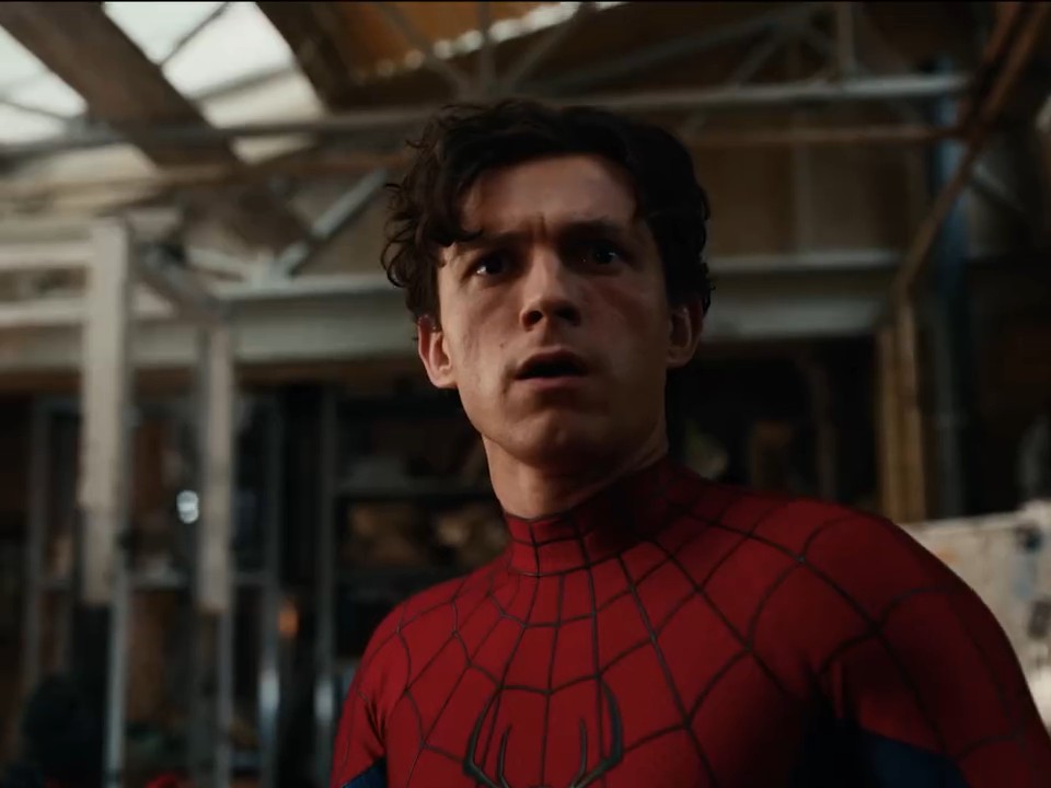 A close-up shot of Tom Holland as Spider-Man in his red and blue suit, looking upward with a concerned expression in what appears to be a warehouse or industrial setting.