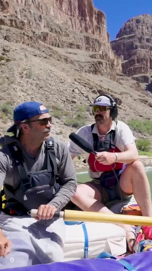 Two men wearing sunglasses and life vests sit in a raft with towering red canyon walls in the background, one holding a microphone while they navigate the Colorado River through the Grand Canyon. The image captures the rugged backcountry adventure described in the post, visually representing the "total backcountry chaos" of their 280-mile journey with a cameraman/mic operator documenting the experience mid-river.