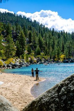 Two people stand at the edge of a clear turquoise alpine lake, surrounded by towering evergreen trees and rocky shorelines, capturing the serene natural beauty that makes Tahoe so special.