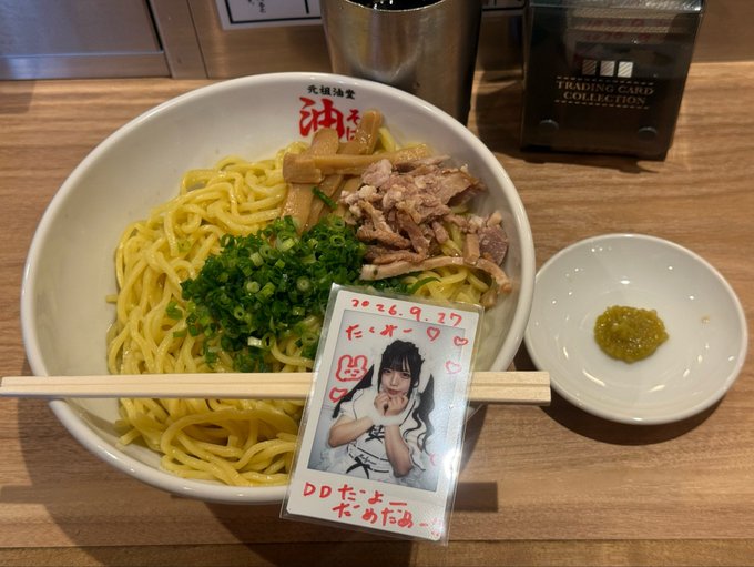A bowl of ramen noodles with green onions and meat is served at a table alongside a small dish of green condiment and a photo card with handwritten text including '2026.9.27' and 'DDたーよー'.