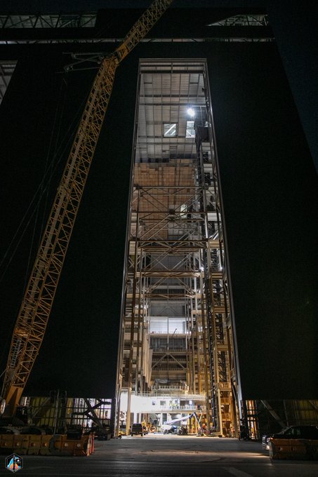 A massive industrial assembly building stands illuminated at night, with a yellow construction crane towering alongside its open facade revealing multi-level scaffolding and structural framework inside. The towering scale and active construction state suggest ongoing work on a large aerospace or heavy industrial project, consistent with a late-September 2026 timeframe.