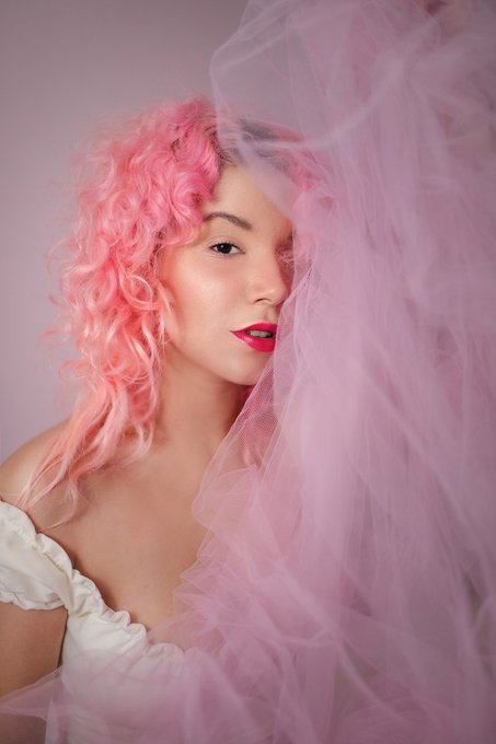A studio portrait of a woman with curly pink hair and bright pink lipstick, partially obscured by flowing pink tulle fabric against a light pink background.