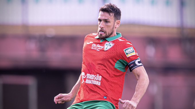 A soccer player with a beard wearing a red and green Maritimo jersey with 'transação máxima' text and a captain's armband, playing in a stadium.