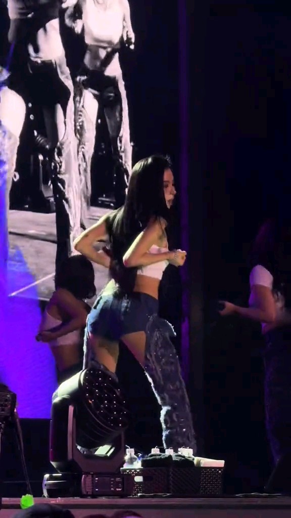A performer with long dark hair dances on stage in a white top and shorts, in front of a large screen and stage lighting.