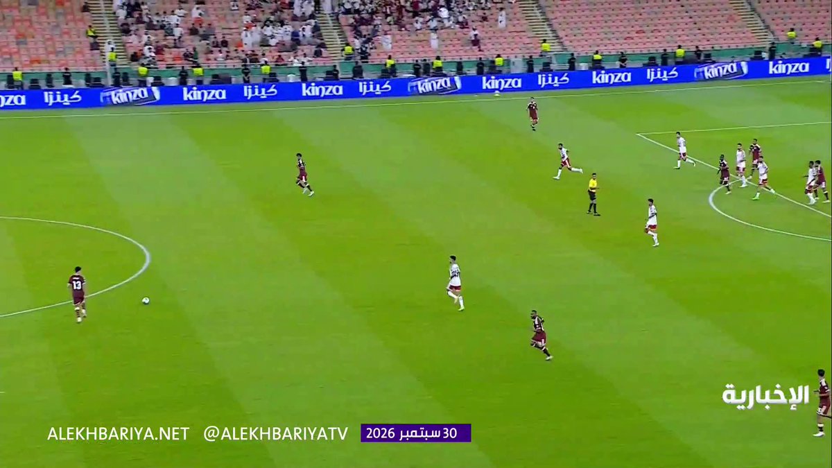 A wide-angle view of a football match in progress on a green pitch, showing players from both teams spread across the field with a referee in yellow nearby, while "Kinza" advertising boards line the stands. The broadcast watermark displays "ALEKHBARIYA.NET @ALEKHBARIYATV" and "30 سبتمبر 2026," corresponding to the post about the UAE vs. Qatar 1-1 match from Gulf Cup 27.