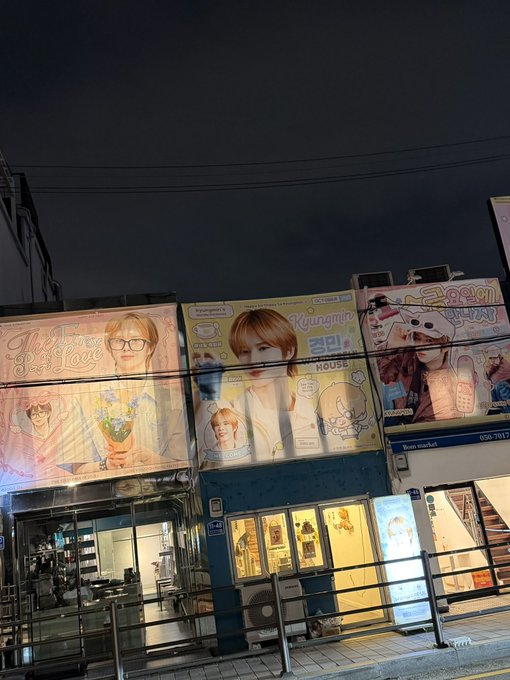 A nighttime street scene showing storefronts with large Korean signage, including billboards featuring a person with glasses advertising 'KYUNGMIN HOUSE' and another sign reading 'Love is always' with 'NO.8' visible on the building.