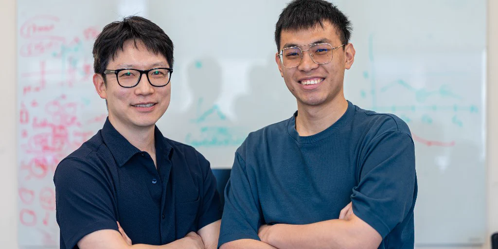 Two individuals are standing in front of a whiteboard with colorful diagrams, with their arms crossed and smiling. They are both wearing glasses. The individual on the left is wearing a dark blue short sleeve button down shirt, and the individual on the right is wearing a lighter blue short sleeve shirt.
