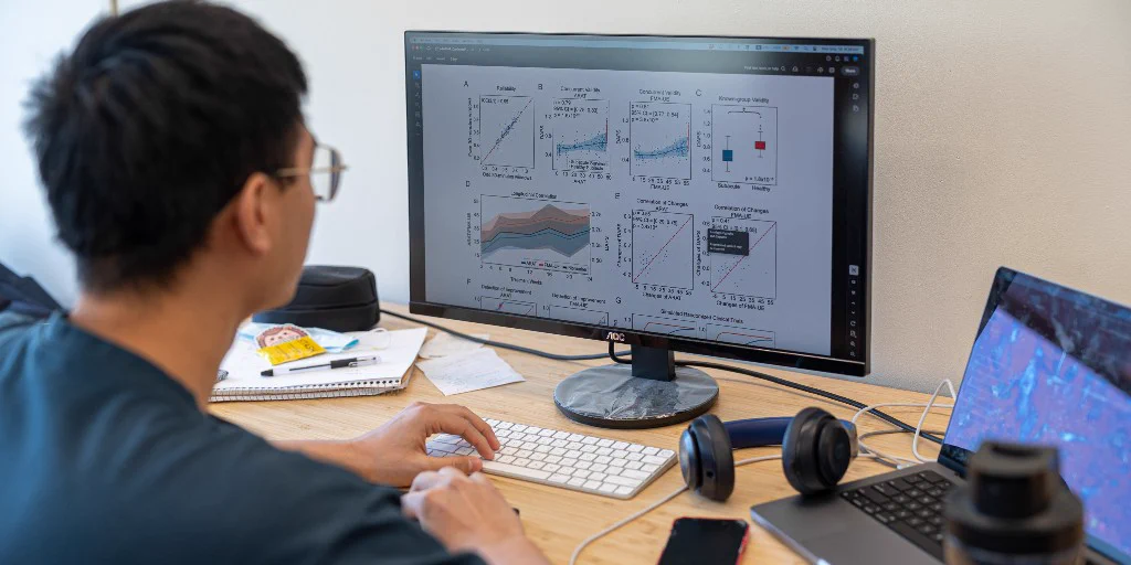 A person is seated at a desk, working on a computer, facing away from the camera. The desktop screen displays graphs and charts. Nearby, there's a laptop with a vibrant screen display. On the desk are headphones, a smartphone, a reusable water bottle, a laptop, a notebook, and other miscellaneous items.
