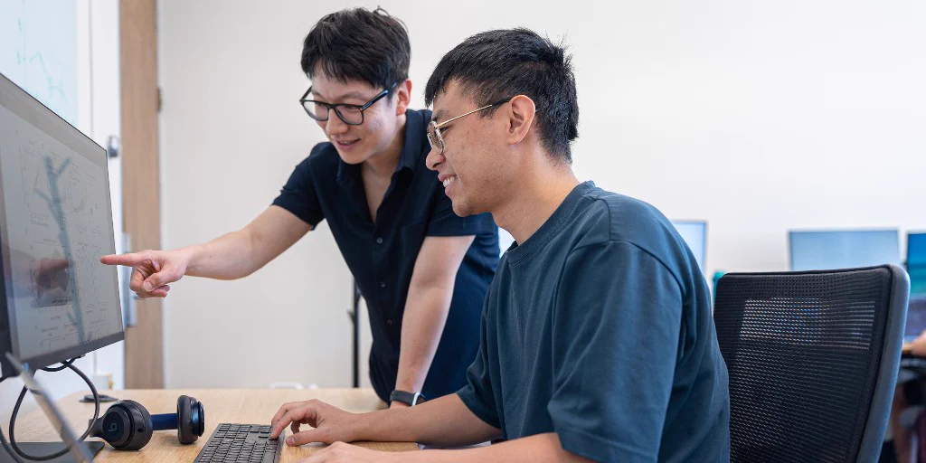 Two people are at a desk with computers. One is seated, working on a computer, while the other stands beside, pointing at the screen. They appear to be in an office or classroom setting, engaging in a collaborative activity. They are smiling.

