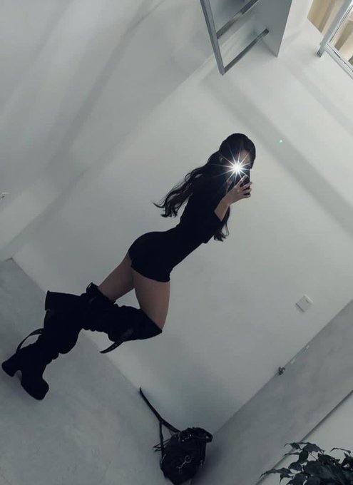 A mirror selfie taken from above showing a woman with long dark hair wearing a black outfit and tall black boots, with a bright camera flash reflecting in the mirror against white walls and floor.