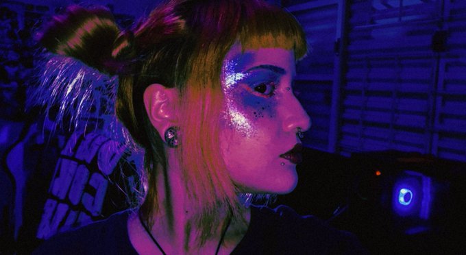 A portrait of a young woman with colorful glitter makeup and space buns, lit with vibrant pink and blue lighting.